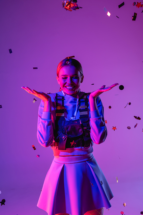 joyful woman in stylish outfit near falling confetti on purple