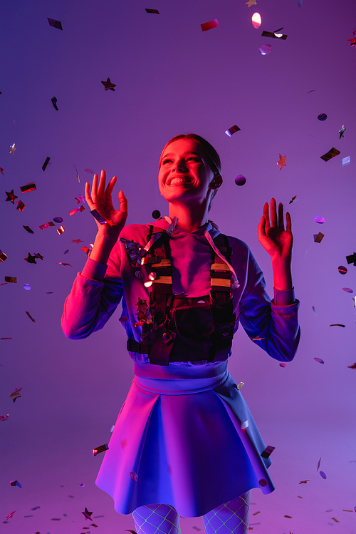 positive woman in stylish outfit near falling confetti on purple