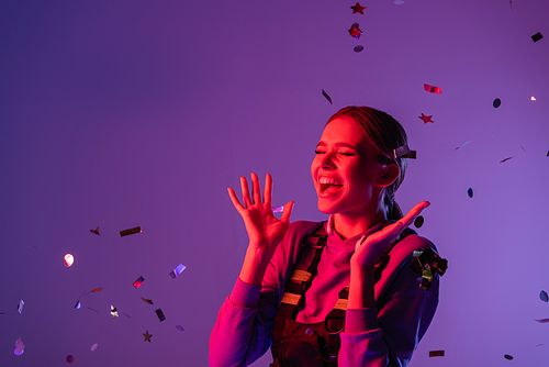 cheerful woman in stylish outfit near falling confetti on purple