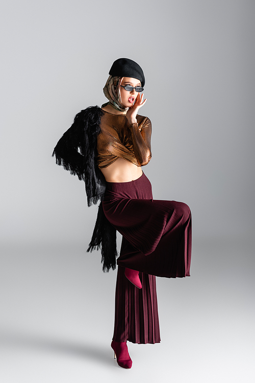 full length of trendy woman in sunglasses and beret posing on grey