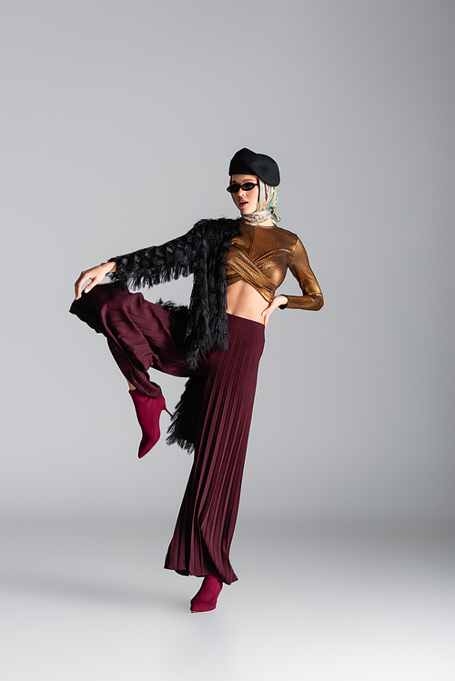 full length of fashionable young woman in sunglasses and beret posing on grey