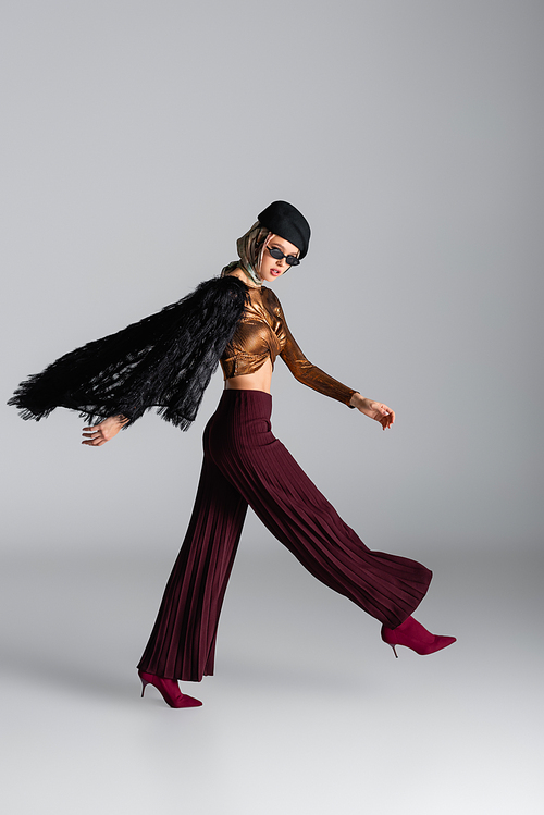 full length of fashionable woman in sunglasses and beret walking on grey