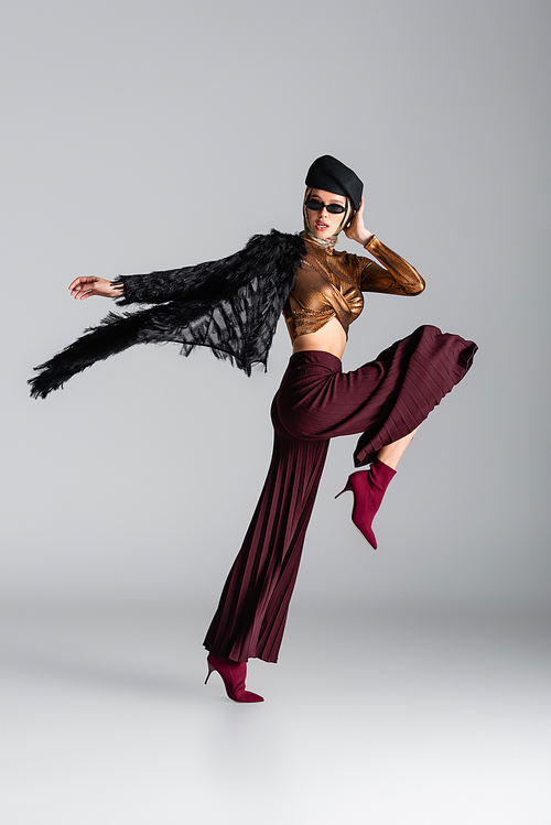 full length of fashionable woman in stylish outfit, sunglasses and beret posing on grey