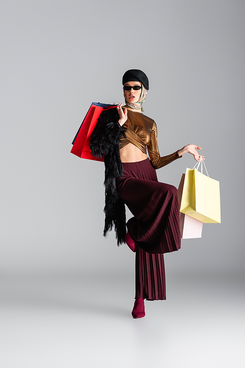 full length of fashionable young woman in sunglasses and beret holding shopping bags   and posing on grey