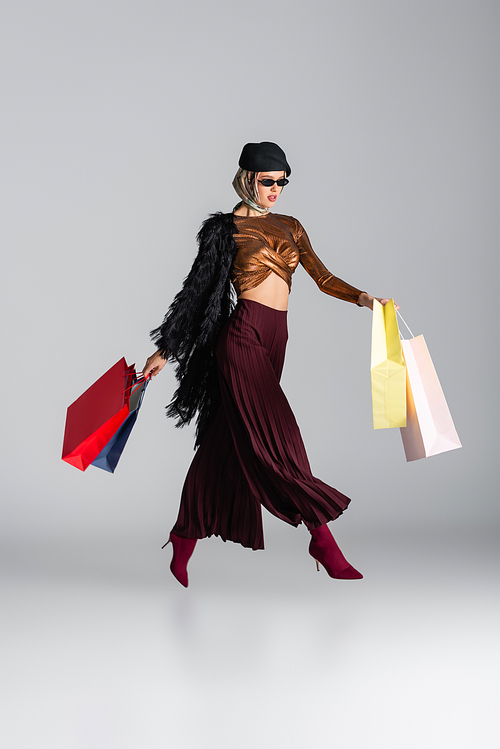 full length of trendy young woman in sunglasses and beret holding shopping bags while levitating on grey