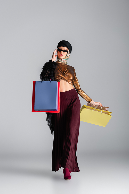 full length of young woman in trendy outfit, sunglasses and beret holding shopping bags while posing on grey