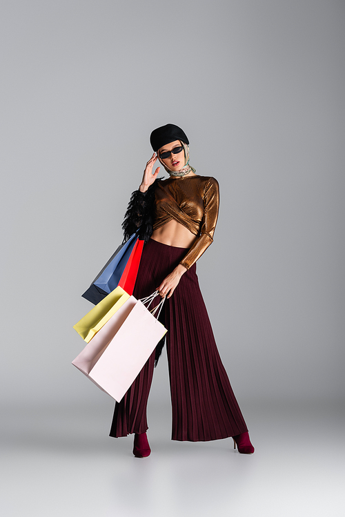 full length of fashionable young woman adjusting sunglasses and holding shopping bags while posing on grey