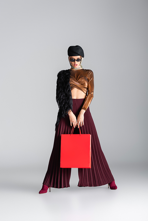 full length of young woman in trendy outfit holding red shopping bag while posing on grey