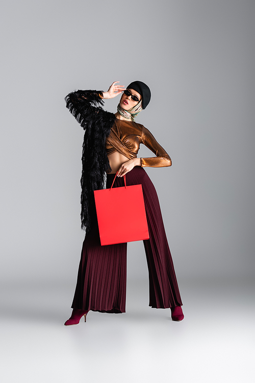 full length of woman in trendy outfit holding red shopping bag while posing on grey