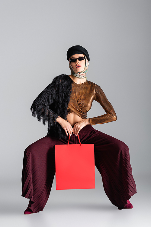 full length of young woman in trendy outfit holding shopping bag while posing on grey