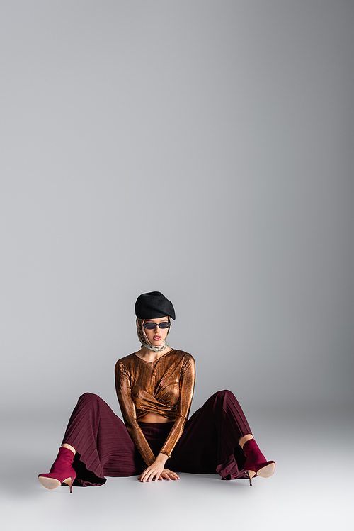 full length of stylish woman in trendy outfit, sunglasses and beret sitting while posing on grey