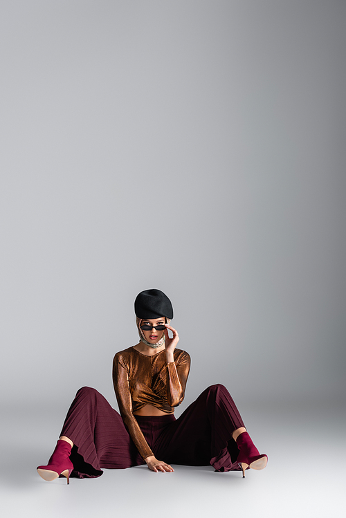 full length of stylish woman in trendy outfit adjusting sunglasses and sitting while posing on grey