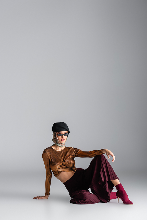 full length of stylish woman in trendy outfit and sunglasses sitting while posing on grey