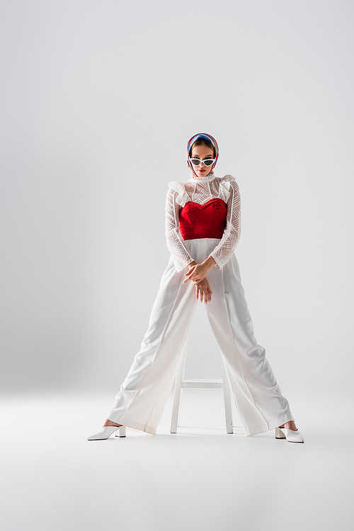 full length of stylish young woman in headscarf and sunglasses sitting on stool and posing on white