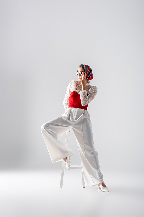 full length of young woman in headscarf and stylish sunglasses sitting on stool and posing on white