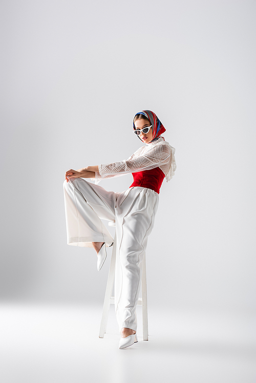 full length of trendy young woman in headscarf and sunglasses posing on stool and  on white