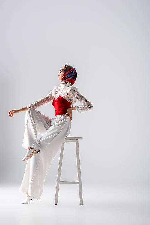 full length of stylish young woman in headscarf and sunglasses sitting with hand on hip on stool and looking away on white