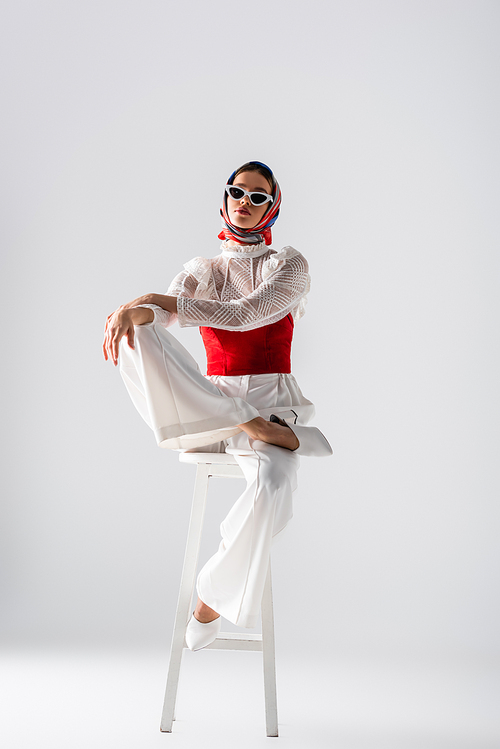 full length of stylish woman in headscarf and sunglasses sitting on stool while posing on white