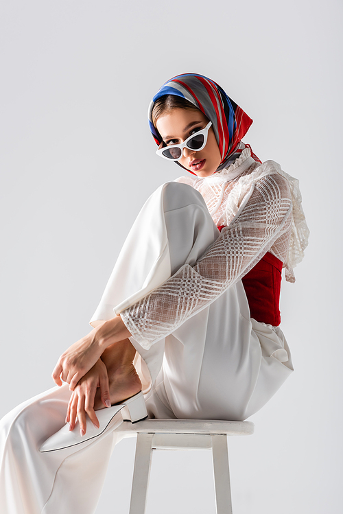 stylish young woman in headscarf and sunglasses sitting on stool while posing isolated on white