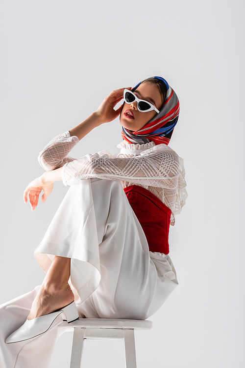 young woman in headscarf and stylish sunglasses sitting on stool while posing isolated on white