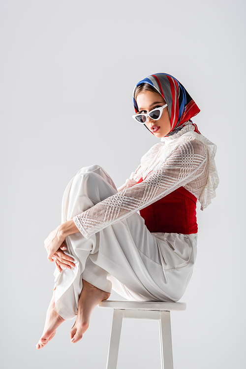 stylish woman in headscarf and sunglasses sitting on stool while posing isolated on white