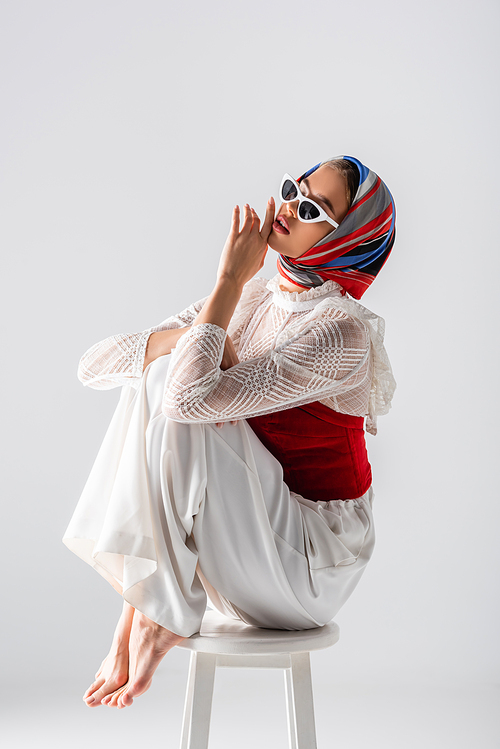 young trendy woman in headscarf and sunglasses sitting on stool while posing isolated on white