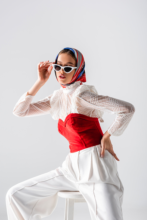 stylish woman in headscarf adjusting sunglasses and sitting on stool with hand on hip isolated on white