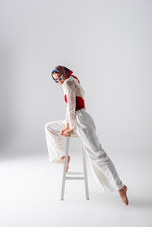 full length of stylish woman in headscarf and sunglasses posing on stool while  on white