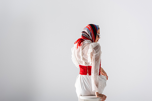 stylish woman in headscarf and sunglasses sitting on stool while looking away on white