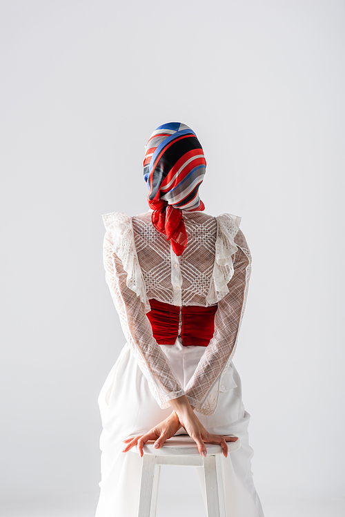 back view of stylish woman in headscarf sitting on stool isolated on white