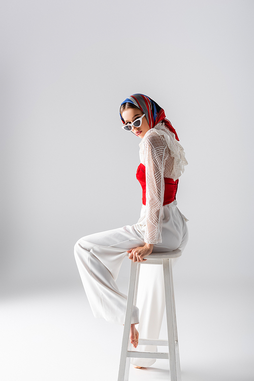 full length of stylish woman in headscarf and sunglasses sitting on stool while  on white