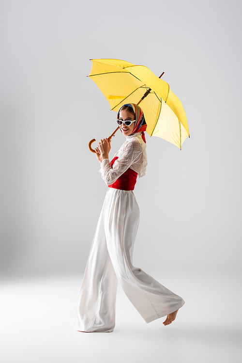 full length of happy woman in headscarf and sunglasses holding yellow umbrella while posing on white