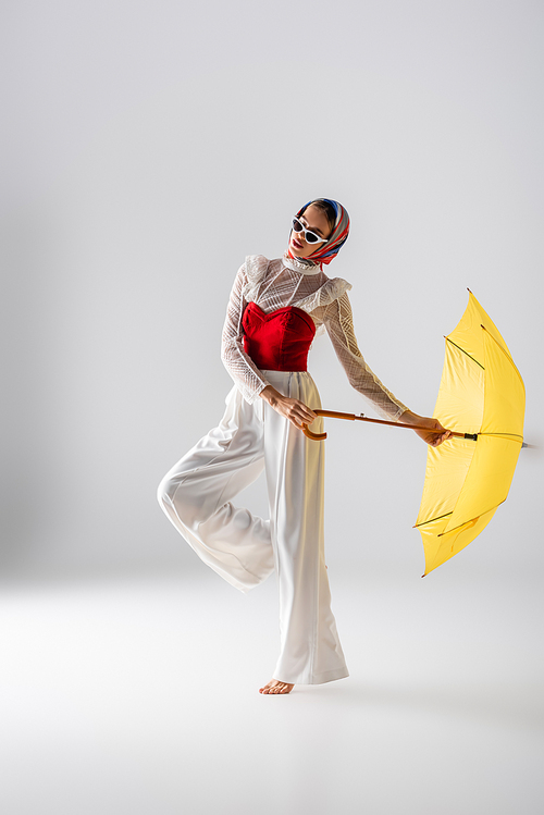 full length of stylish woman in headscarf and sunglasses holding yellow umbrella while posing on white