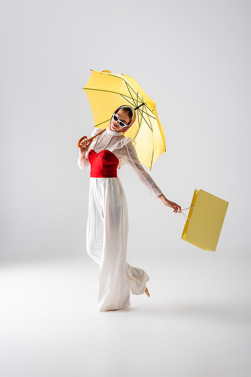 full length of happy woman in headscarf and sunglasses holding yellow umbrella and shopping bag while posing on white