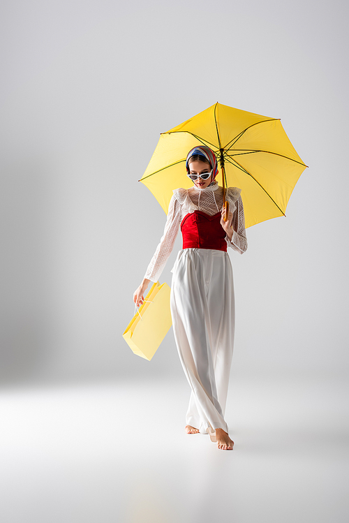 full length of stylish woman in headscarf and sunglasses holding yellow umbrella and shopping bag while posing on white