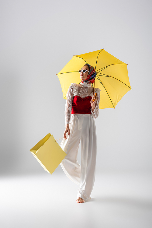 full length of young woman in headscarf and sunglasses holding yellow umbrella and shopping bag while posing on white