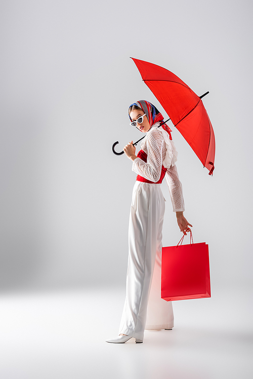 full length of young woman in headscarf and sunglasses holding red umbrella and shopping bag while posing on white