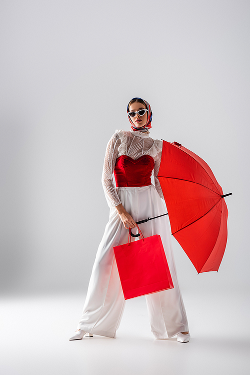 full length of fashionable woman in headscarf and sunglasses holding red umbrella and shopping bag while posing on white