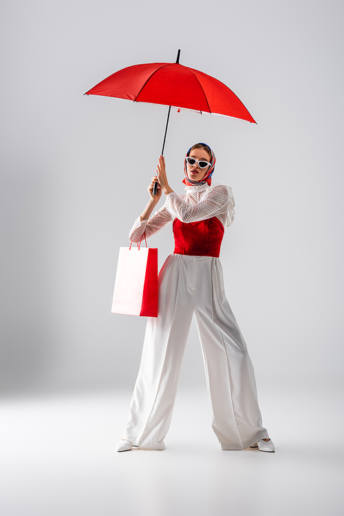 full length of stylish woman in headscarf and sunglasses holding shopping bag while standing under umbrella on white