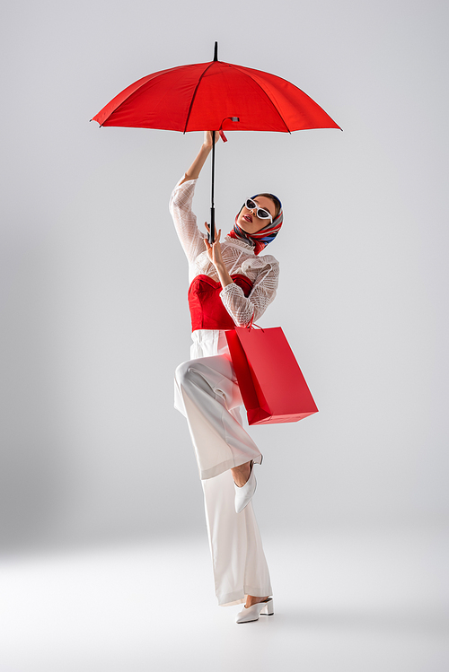 full length of young woman in headscarf and stylish sunglasses holding red umbrella and shopping bag while posing on white