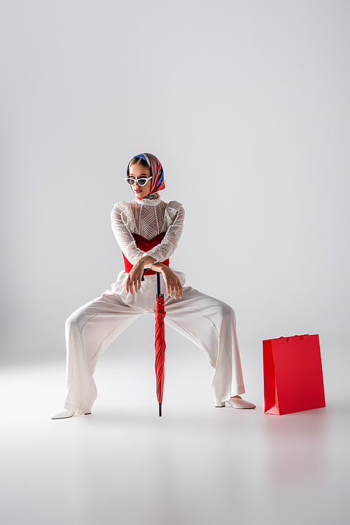 full length of stylish woman in headscarf and sunglasses holding red umbrella near shopping bag while posing on white