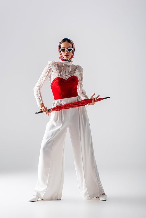 full length of stylish woman in headscarf and sunglasses holding red umbrella while posing on white