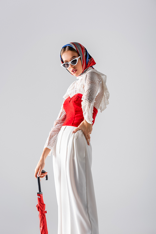 stylish woman in headscarf and sunglasses holding red umbrella while posing on white