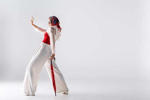 full length of stylish woman in headscarf and sunglasses holding red umbrella while gesturing  on white