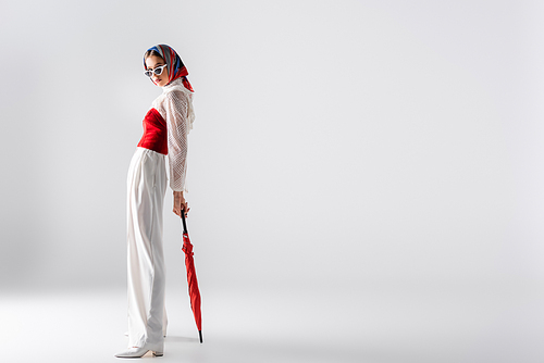 full length of young woman in headscarf and sunglasses holding red umbrella while posing on white