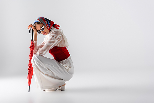 full length of stylish woman in headscarf and sunglasses holding red umbrella while sitting on white