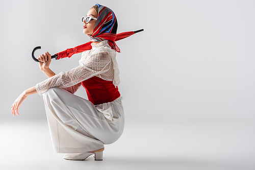 full length of young woman in headscarf and sunglasses holding red umbrella while sitting on white