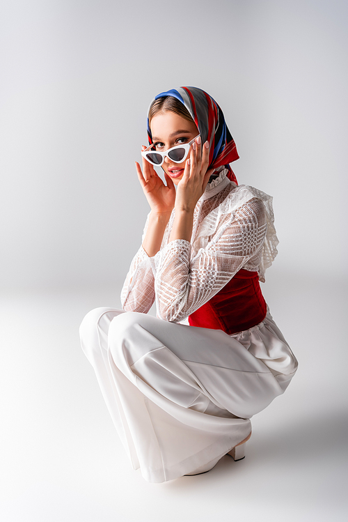 stylish woman in headscarf adjusting sunglasses while sitting on white