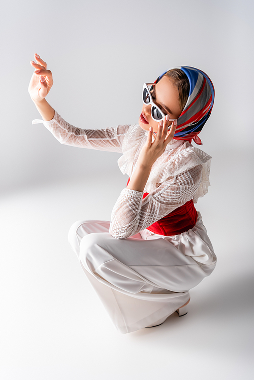 stylish woman in headscarf adjusting sunglasses, gesturing and sitting on white