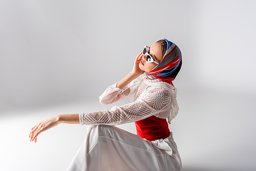 trendy woman in headscarf adjusting sunglasses and sitting on white